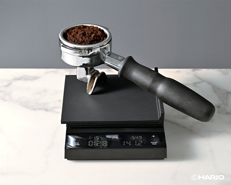 square silicone mat on top of a digital scale weighing a espresso portafilter with coffee ground. on a marble table and gray background.