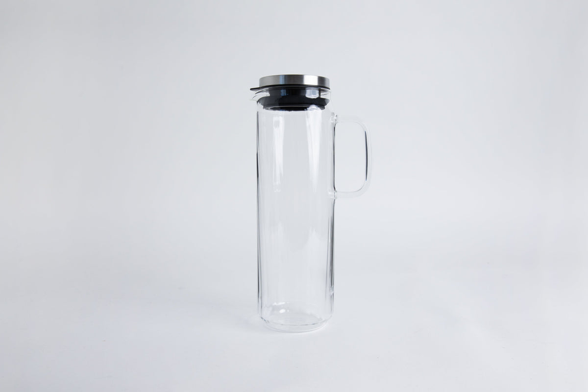 A tall clear glass pitcher with glass handle with metal lid and black gasket. seton a white background