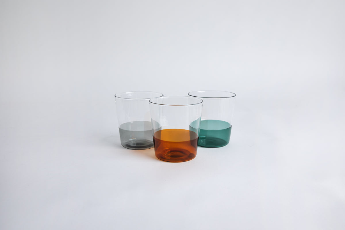 3 dual colored clear glass cup side by side, one cup top half is clear and second half colored amber, one cup top half is clear and second half colored gray, one cup top half is clear and second half colored green. set on a white background