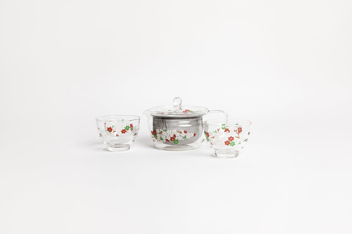 All glass tapered tea pot with flat bottom, all glass lid and handle, and metal mesh filter basket insert. decorated glass with a minimal flower design with match small all glass tea cups with flower design. set on a white background.
