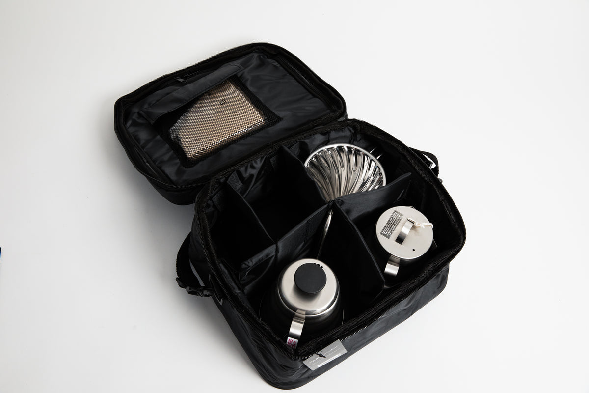 black carrier bag with compartments with stainless steel kettle, metal cone coffee dripper with a olive green base on top of a stainless steel coffee sever pot with stainless steel lid and coffee filters on a white background.