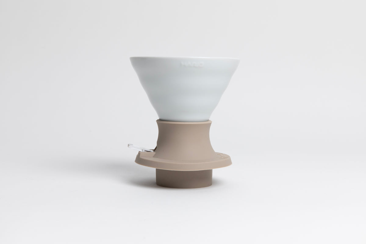 Cone-shaped matte white ceramic dripper seated in a light tan silicone base with clear plastic push switch on a white background.