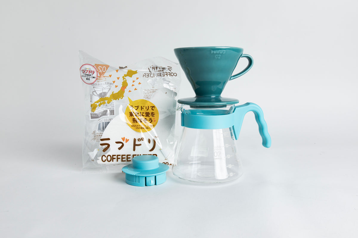 Teal HARIO 02 cone dripper on a glass carafe with a light teal handle, shown with paper filters and a matching teal lid on a light gray background.