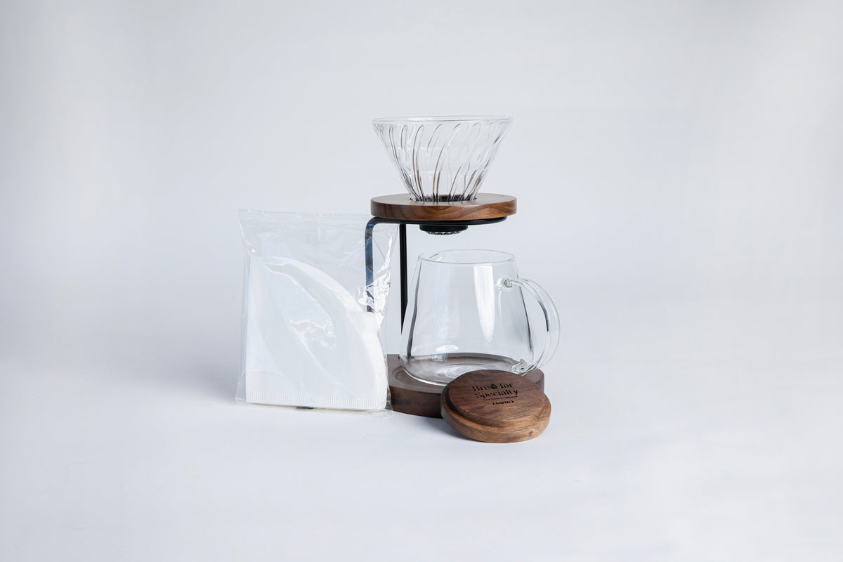 Clear all glass cone shaped coffee dripper with ribs, sitting in a round walnut wood base. Dripper is on a on metal and walnut wood hanging stand underneath a a tulip shape glass decanter with glass handle. With a walnut lid for the decanter and a triangular shaped filters in a cellophane bag. set on a white background.