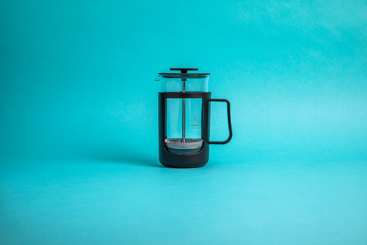 Coffee press featuring a glass pitcher with fluted spout, stainless steel mesh filter with pole, and a black plastic lid, knob, holder, and handle. Set against a blue background.