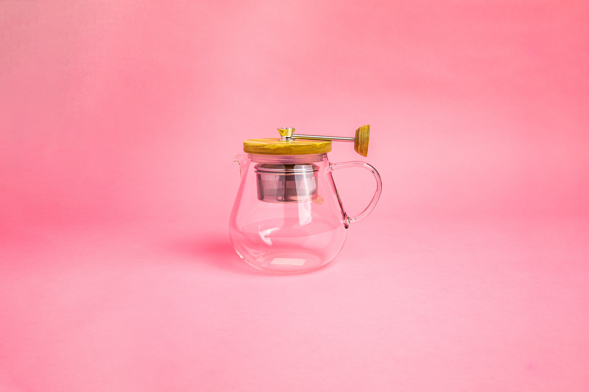 Glass teapot with glass handle. Olive wood lid and knob. Steel rod with a steel tea filter basket. set on pink gradient background
