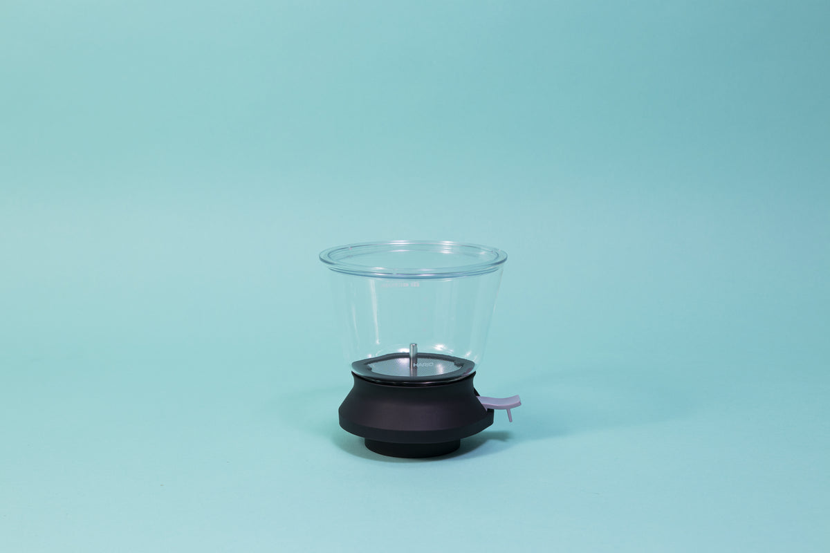 Cylindrical glass tea brewer and metal mesh strainer with clear plastic lid atop a black rubber base with plastic lever