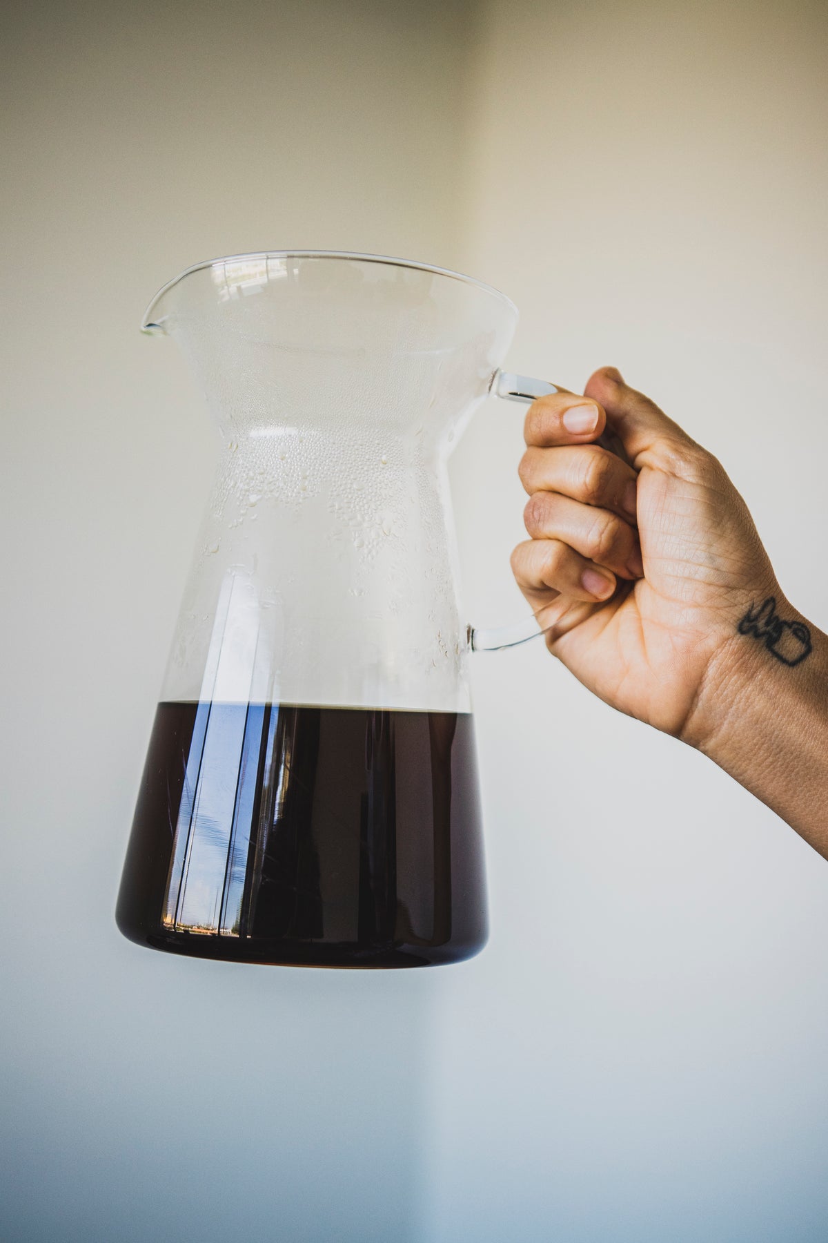 Model's hand holding a tapered clear glass server with fluted spout, all glass handle, and coffee inside.