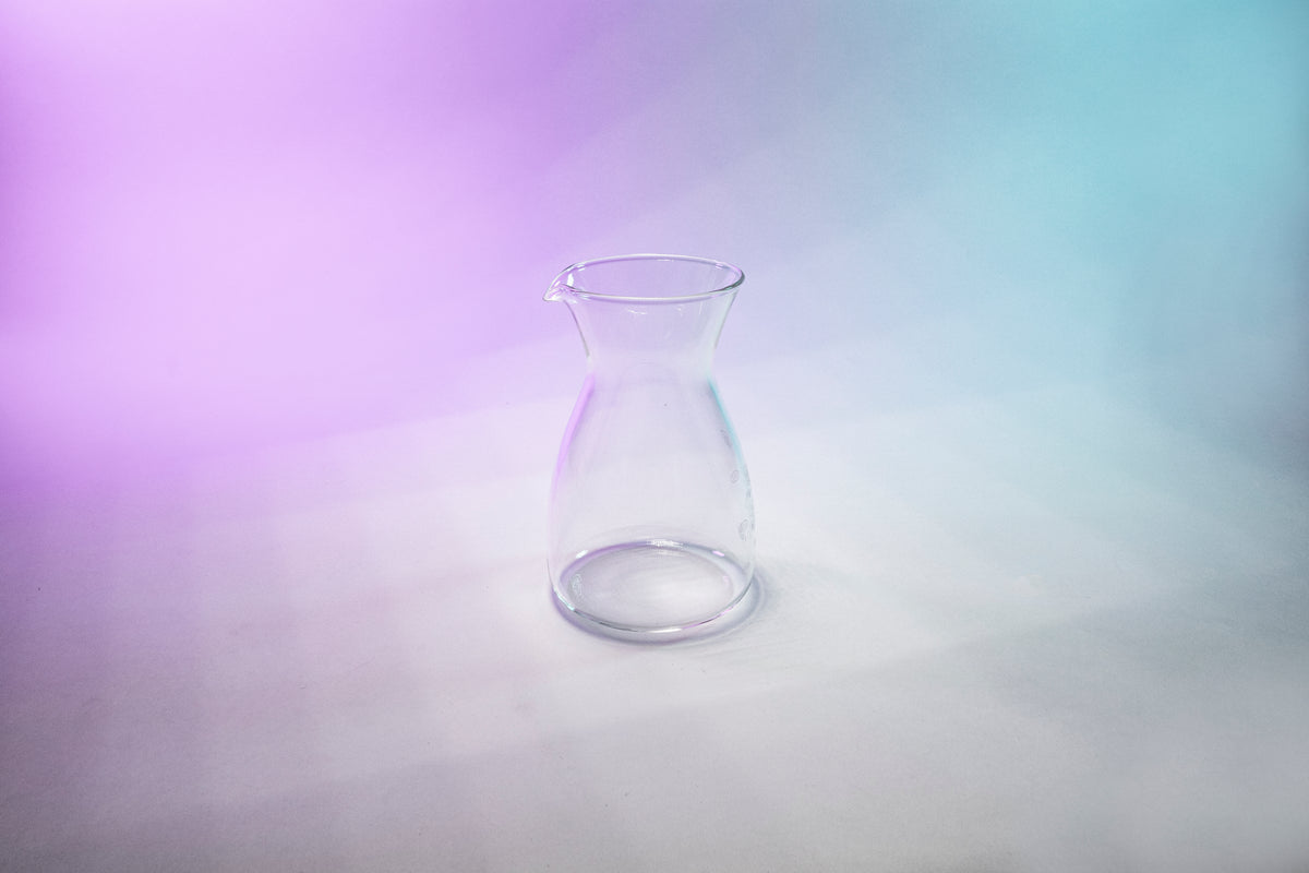 Tapered glass coffee decanter with flared spout against a purple, grey, and blue background.