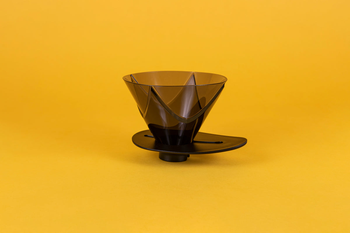 Translucent black plastic cone shaped dripper with bezier ridges and oval shaped black plastic base on a yellow background.