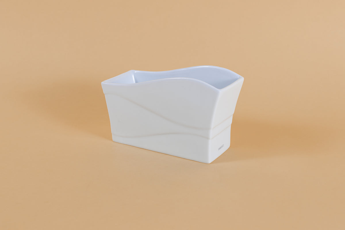 Rectangular all white ceramic vessel with "wave" design along side and top on orange backdrop.
