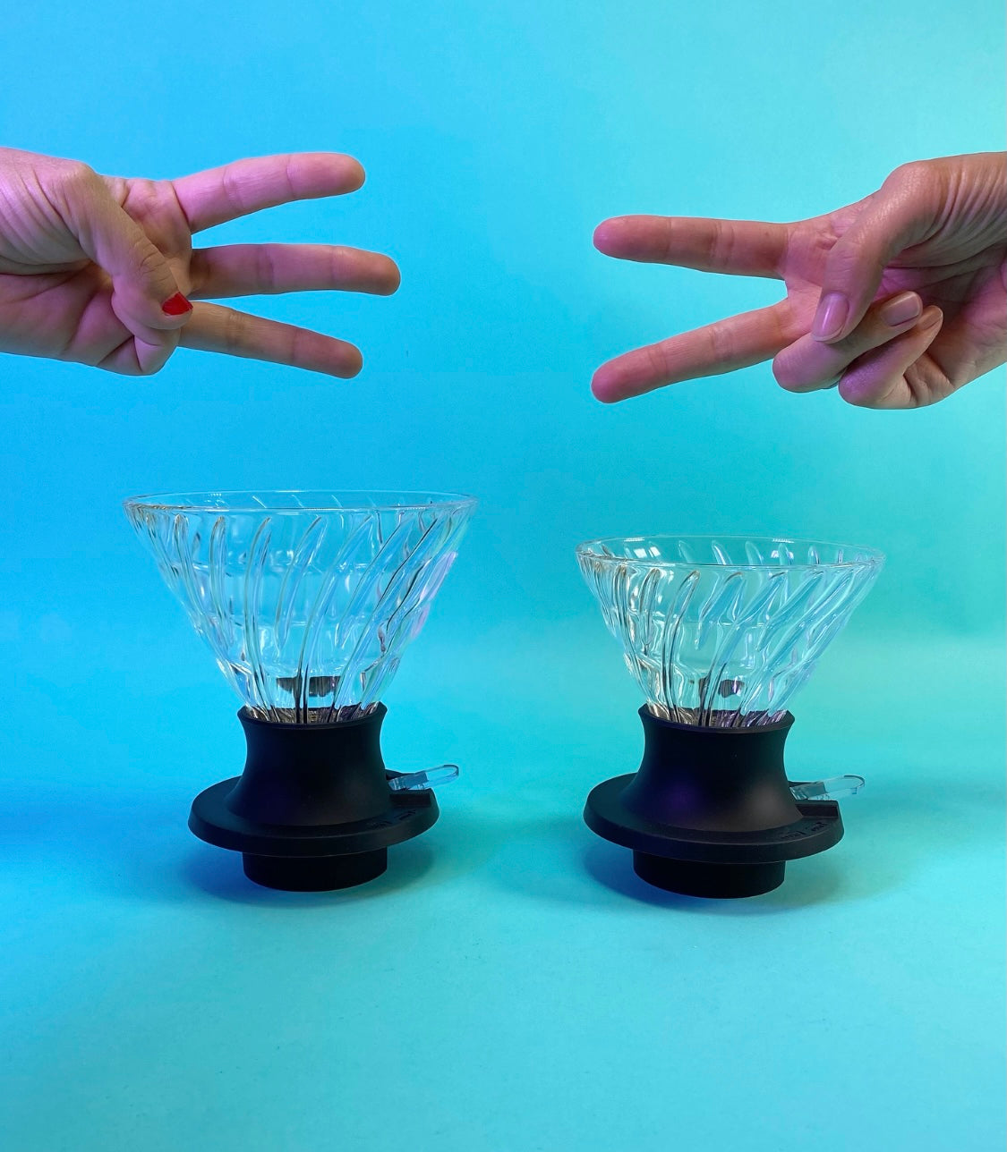 Large cone-shaped glass dripper with black silicone base with clear plastic switch, and a hand holding three fingers above to indicate size, next to a medium cone-shaped glass dripper with black silicone base with clear plastic switch, and a hand holding two fingers above to indicate size, on a light blue background.