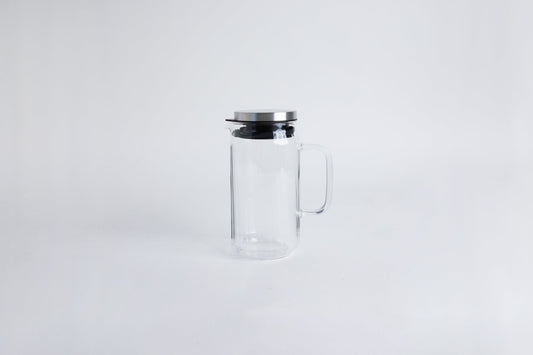 Clear glass pitcher with glass handle with metal lid and black gasket. Set on a white background