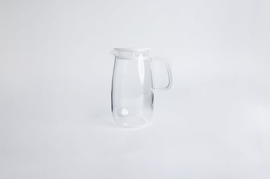 Round clear glass pitcher with glass handle and white lid set on a white background