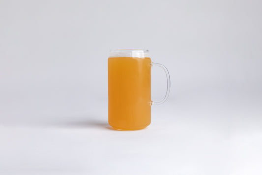 A tall clear mug  with glass handle filled with orange liquid with a foam top. set on a white background