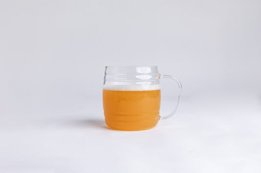 A clear ribbed glass mug with a yellow liquid and white foam top set on a light gray background