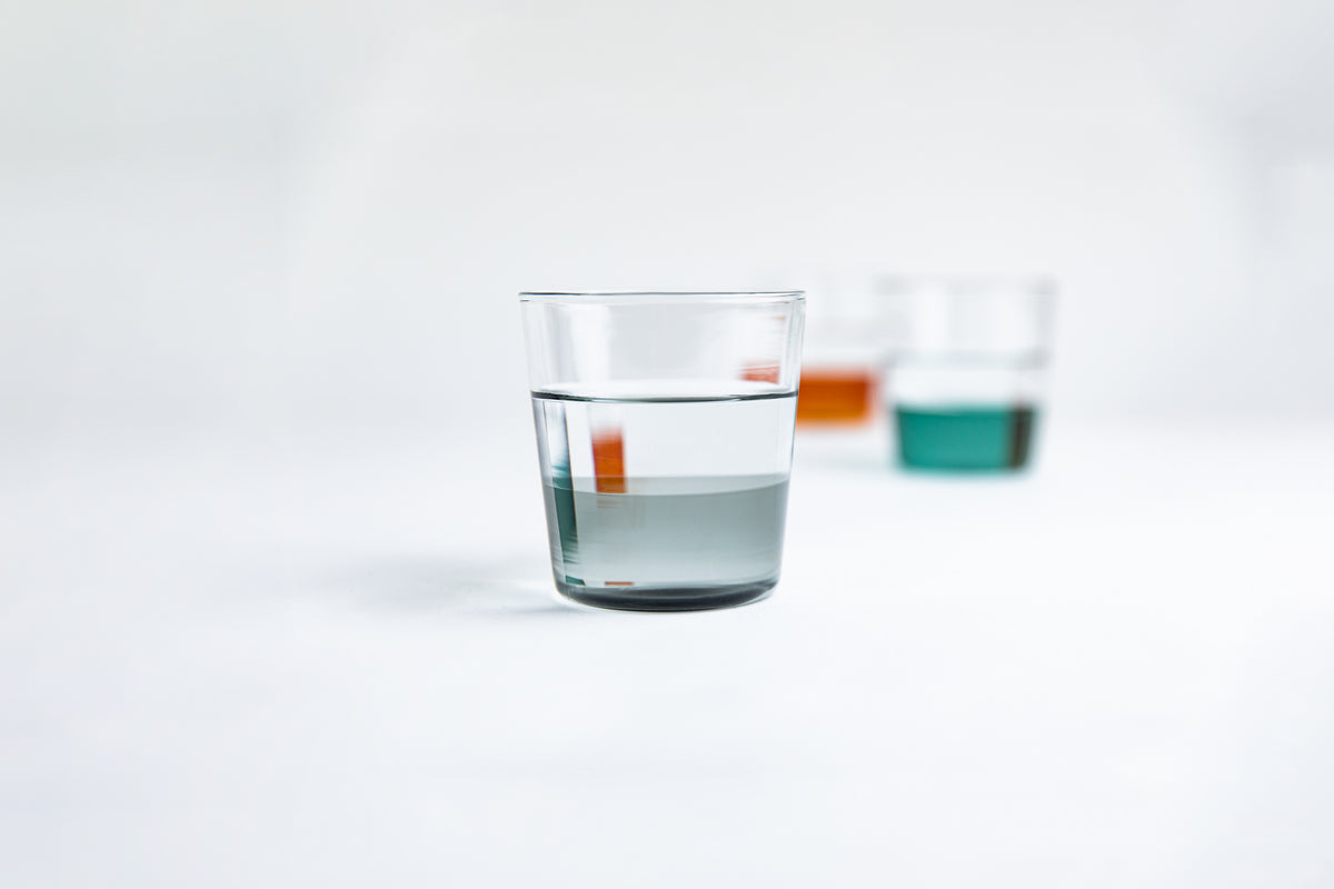 Dual colored clear glass cup, top  half is clear and second half colored gray filled with water. Foreground blur photo of dual colored clear glass cup, top  half is clear and second half colored green and other cup is amber set on a white background 