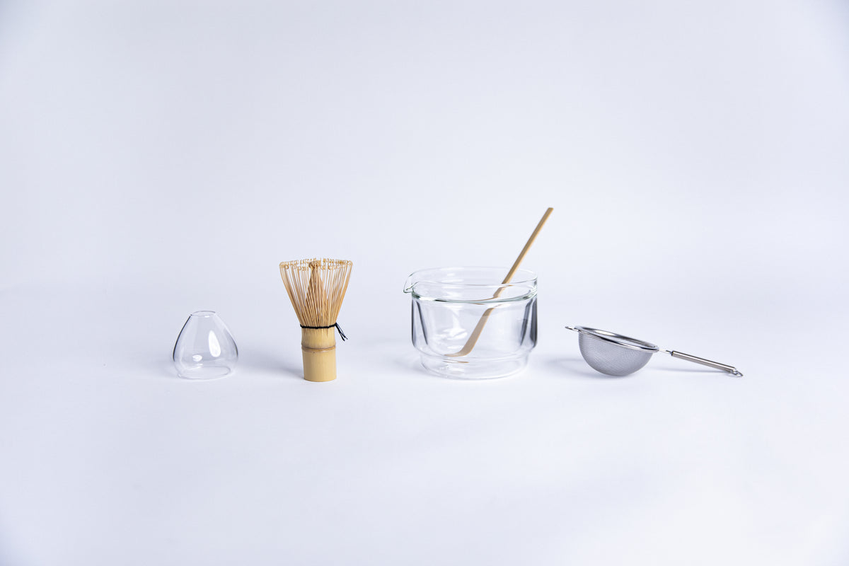 Matcha tea-making tools including a bamboo whisk, clear glass dome shape bamboo whisk holder, Double-walled glass cup, stick tool and metal sieve. set on a white background.