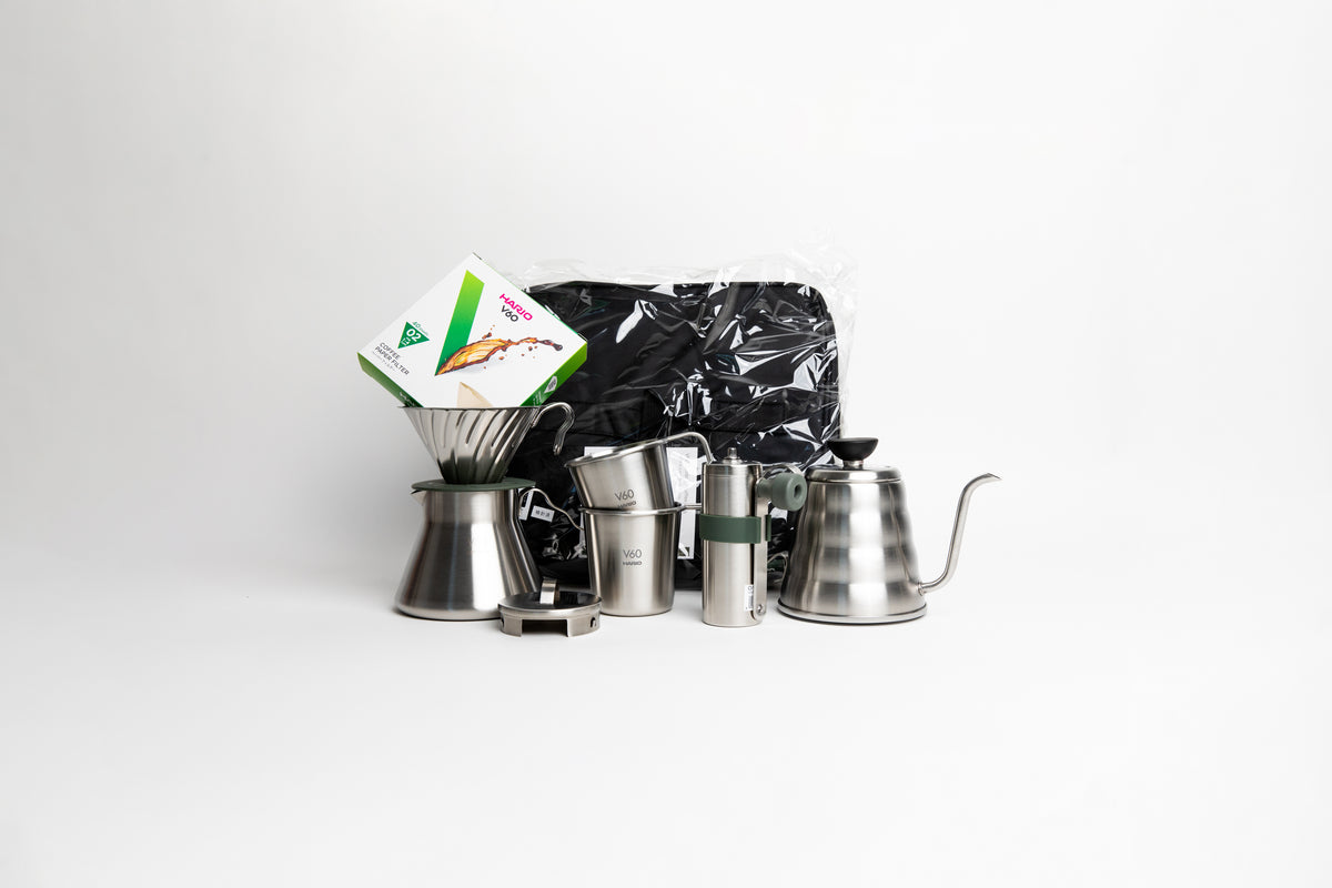 stainless steel kettle, metal cone coffee dripper with a olive green base on top of a stainless steel coffee sever pot, stainless steel lid, with metal coffee hand grinder and two metal cups mugs, box of filters and a black bag. on a white background