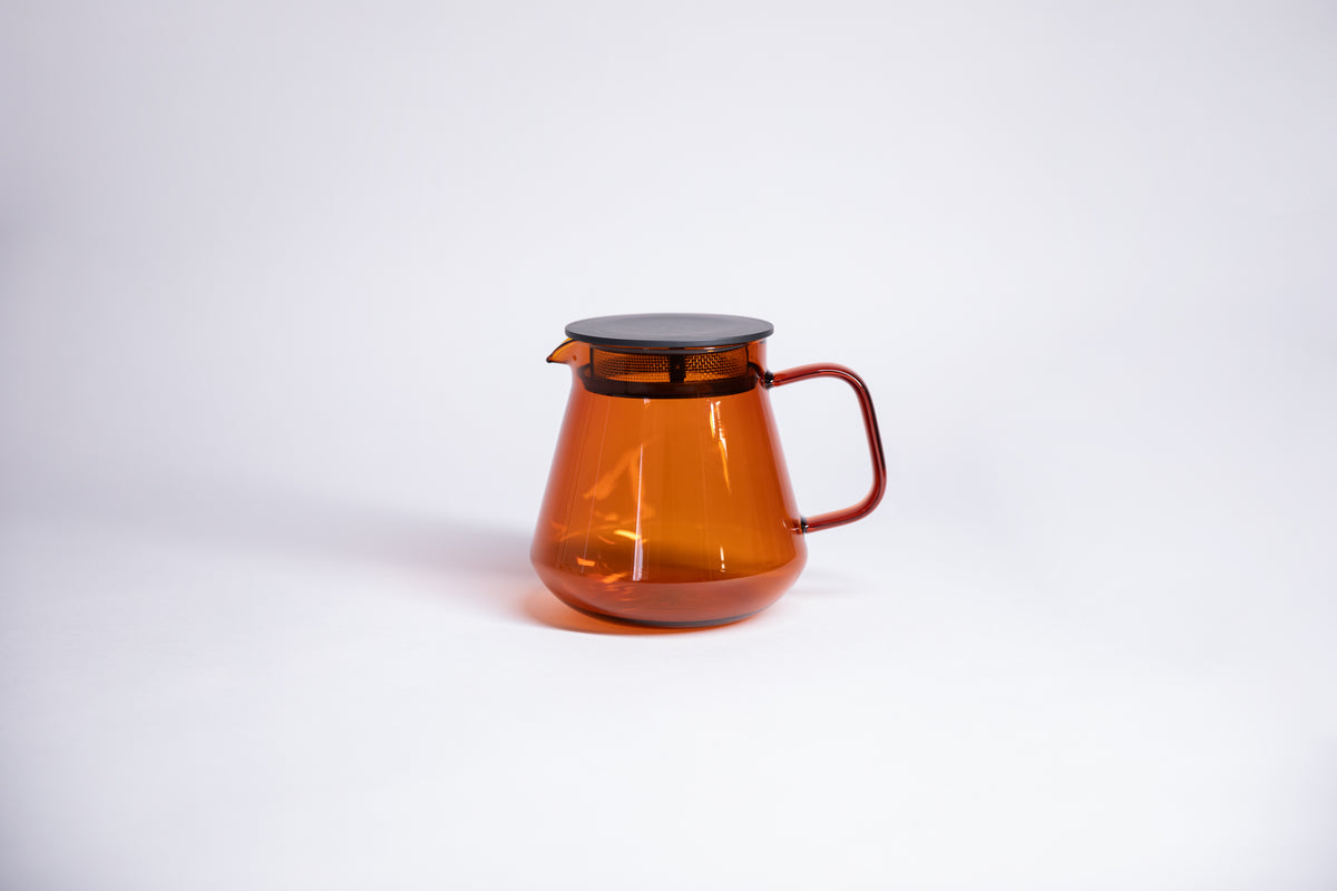 Transparent clear amber colored tulip shaped tea or coffee server with glass handle and fluted spout and black lid with a built in strainer. Set on white background