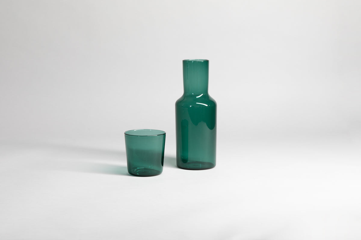 Transparent green glass carafe with matching glass cup set on the side. Set on a light gray background