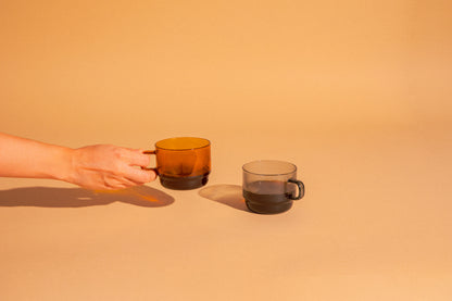 Hand holding amber glass mug with coffee. Transparent gray glass mug with coffee set on a light terracotta background