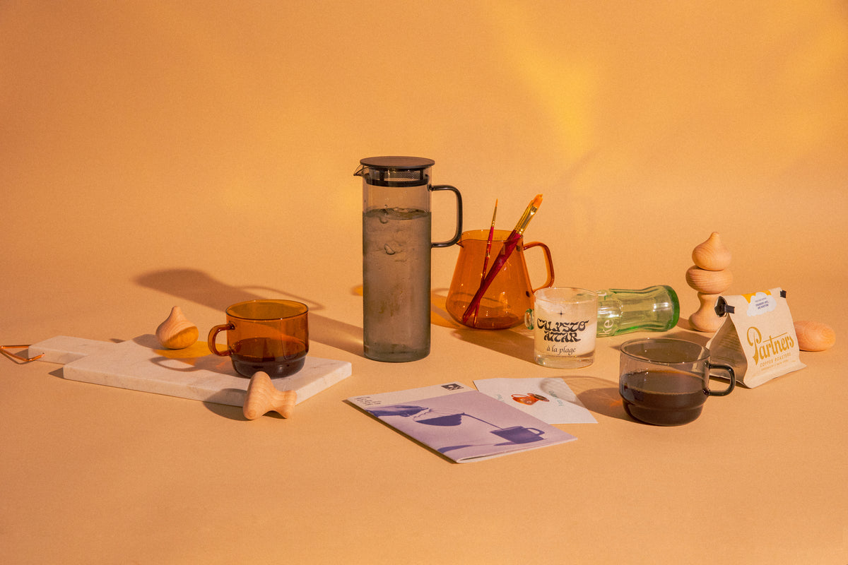 Transparent glass amber mug filled with coffee and transparent glass amber tea/coffee server with 2 decorative paint brushes. Transpartment cylindrical grey pitcher with handle filled with water. Transparent grey glass mug with coffee. Objects surrounded with decorative art pieces. set on a orange background
