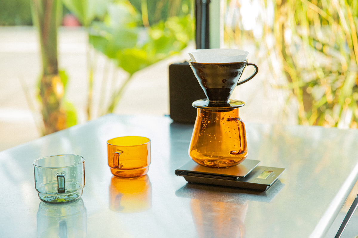  gray and amber colored transparent glass mugs. Black HARIO V60 top of amber glass transparent server with handle sitting on a black pour over scale.