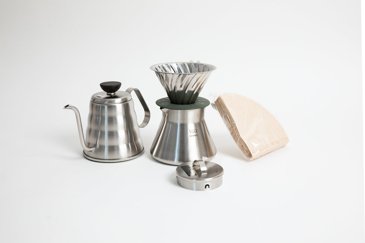 stainless steel kettle, metal cone coffee dripper with a olive green base on top of a stainless steel coffee sever pot, stainless steel lid and coffee filters on a white background. 