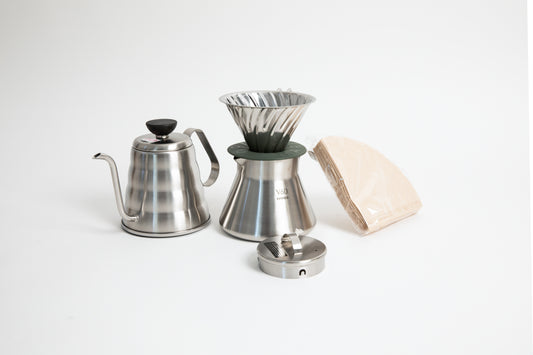 stainless steel kettle, metal cone coffee dripper with a olive green base on top of a stainless steel coffee sever pot, stainless steel lid and coffee filters on a white background. 