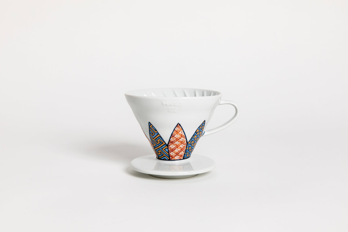 White cone-shaped ceramic coffee dripper with handle and round base, featuring Shonzui (祥瑞) pattern with auspicious symbols like pine, bamboo, and plum blossoms on a geometric tortoise-shell design. set on a white background.