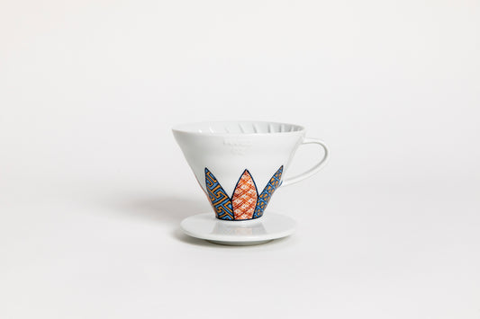 White cone-shaped ceramic coffee dripper with handle and round base, featuring Shonzui (祥瑞) pattern with auspicious symbols like pine, bamboo, and plum blossoms on a geometric tortoise-shell design. set on a white background.