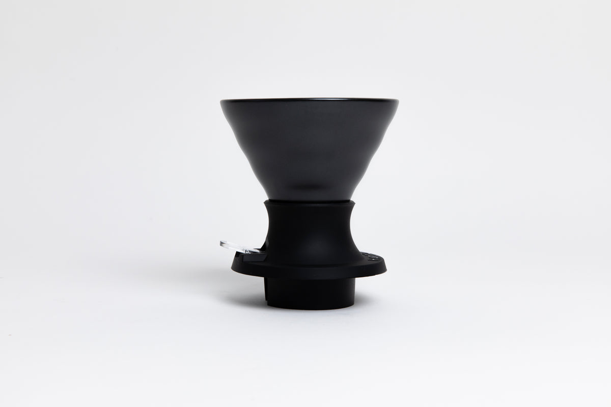 Cone-shaped matte black ceramic dripper seated in a black silicone base with clear plastic push switch on a white background.