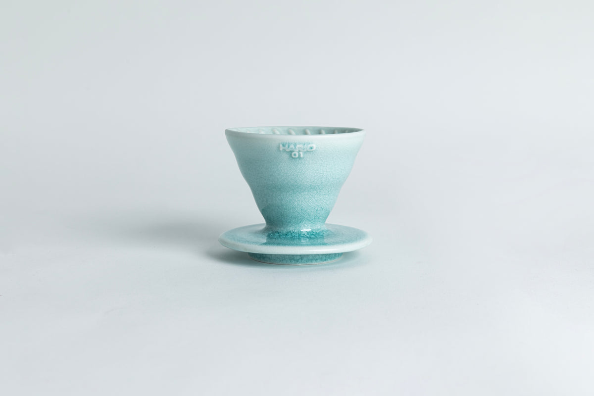 A ceramic pour-over cone with a saucer base featuring a gradient crackle glaze from light teal to teal. The text 'HARIO 01' is displayed on the cone. Set against a light gray background.