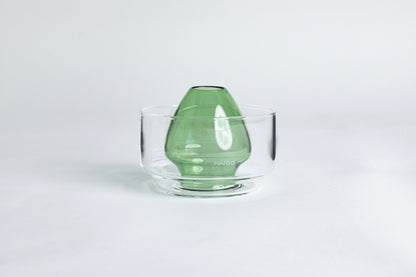 Dome-shaped green clear glass Matcha whisk holder with a smooth, tapered form, placed inside a clear glass base. Displayed on a white background with 'Hario' branding