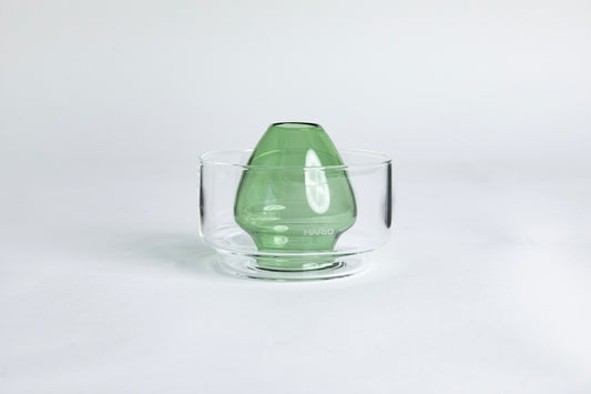Dome-shaped green clear glass Matcha whisk holder with a smooth, tapered form, placed inside a clear glass base. Displayed on a white background with 'Hario' branding
