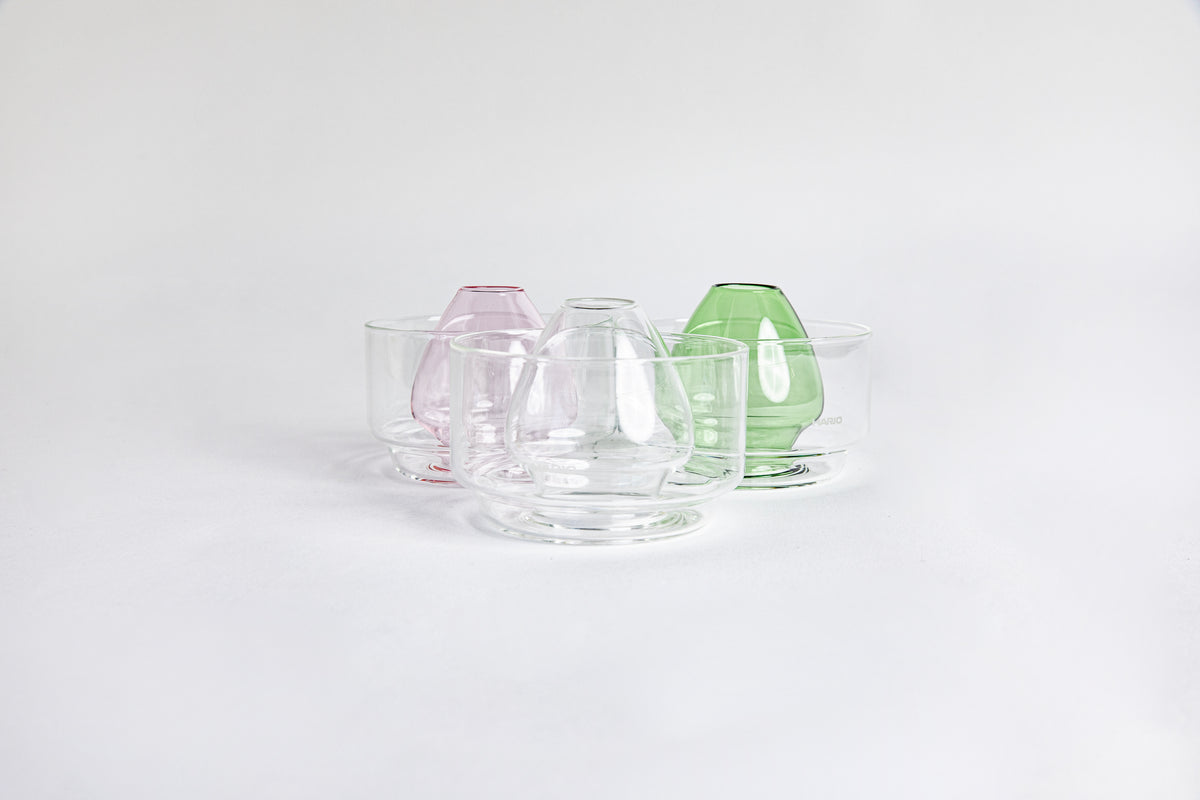 Three side by side images of a dome-shaped in the colors clear, pink and green clear glass matcha whisk holder with a smooth, tapered form, placed inside a clear glass base. Displayed on a white background with 'Hario' branding