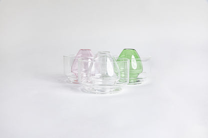 Three side by side images of a dome-shaped in the colors clear, pink and green clear glass matcha whisk holder with a smooth, tapered form, placed inside a clear glass base. Displayed on a white background with 'Hario' branding