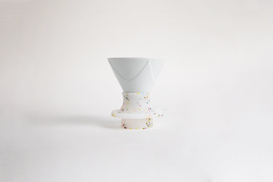 White ceramic cone-shaped dripper with flowing Bezier-style ridges, set in a white silicone base that features rainbow speckles. A clear plastic switch. set on a white background.
