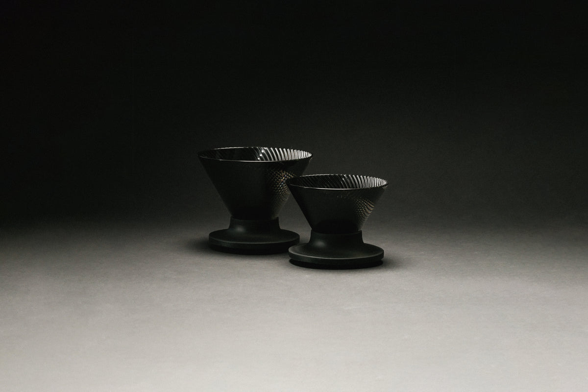 2 black, cone shaped coffee drippers on a black and grey background. 