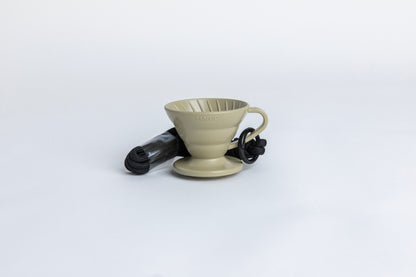 khaki cone shaped plastic coffee dripper  cup with handle and round base.  with a black rope tied on the handle Set on white background