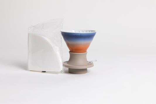 A ceramic HARIO cone-shaped dripper featuring a gradient sunset color scheme of orange, white, and blue, with a white interior. It sits in a gray mauve silicone base with a clear plastic switch. Next to it is a cellophane bag containing triangular paper filters, against a white background.
