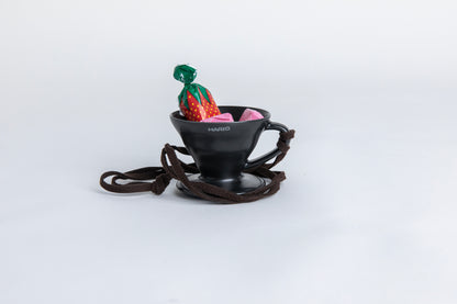 matte black cone shaped ceramic coffee dripper  cup with handle and round base.  with a brown rope tied on the handle. with  small pieces of candy inside showcasing the size of the small cup Set on white background