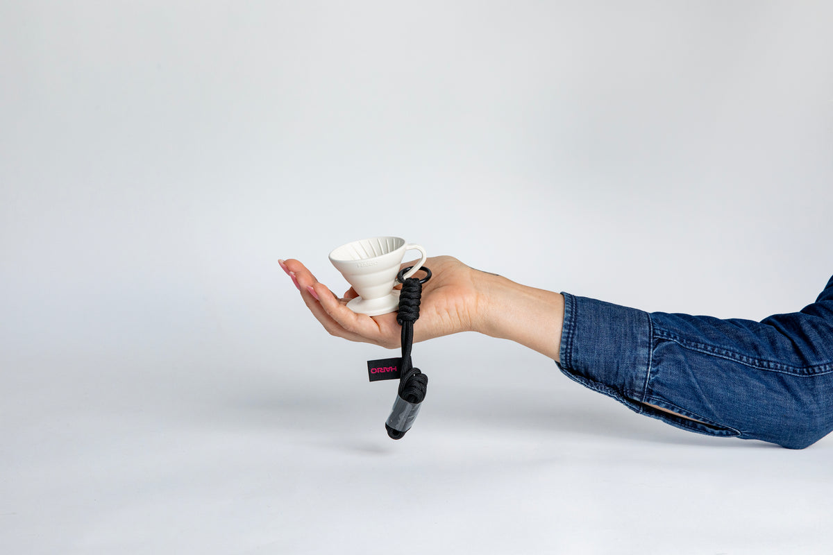 hand holding the white black cone shaped ceramic coffee dripper cup with handle and round base. with a brown rope tied on the handle, showcasing the size of the small cup can fit in the palm of a  hand. Set on white background