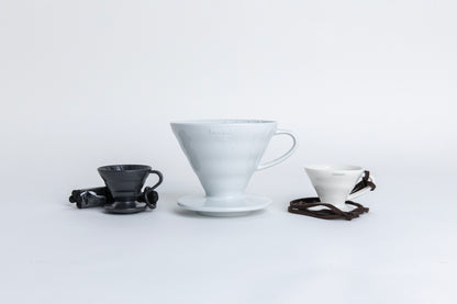 Three cone-shaped coffee drippers with handles and round bases, arranged side by side on a white background. The first is a mini matte black dripper with a brown rope tied around the handle. Next to it is a standard 02-size ceramic dripper in a classic finish, and beside that, a mini white ceramic dripper with the same cone shape and design. set one a white background.