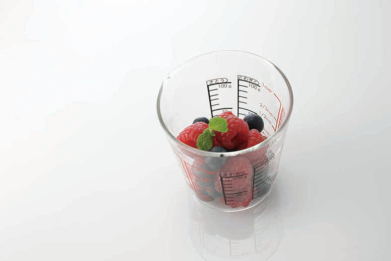 Glass measuring cup on white, reflective background with measurements in red and black on the inside and outside of the glass and partly filled with raspberries and blueberries.