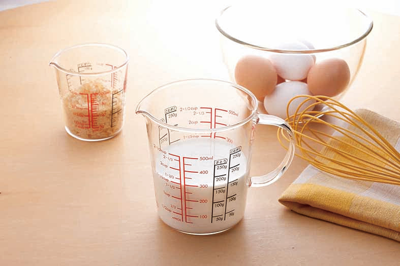 Glass measuring cup with spout, full glass handle, measurements in red and black on the inside and out, and filled with milk on a table setting with a small glass measuring cup with measurments in red and black on the inside and outside of the glass with bread crumbs inside. A yellow whisk on a folded yellow, white and beige checkered hand towel and a glass mixing bowl filled with brown and white eggs.