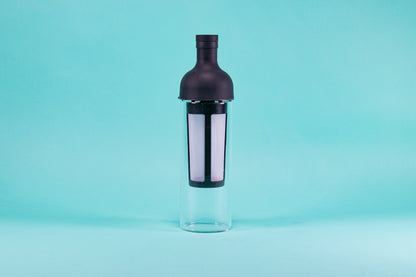 Tall glass container and white nylon mesh coffee filter, with black rubber wine bottle shaped top on a blue background.