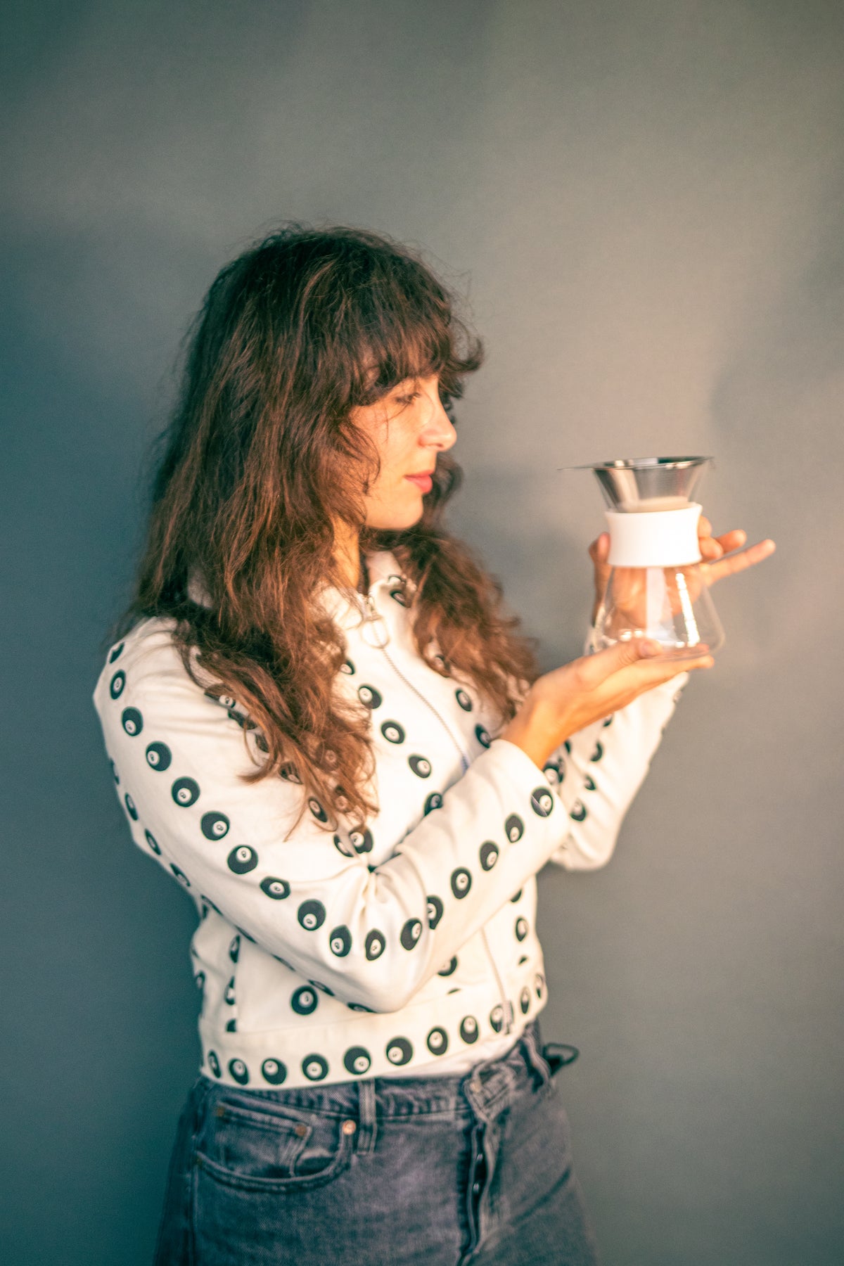Woman holding a clear glass server with white silicone collar and stainless steel mesh filter against a grey background.