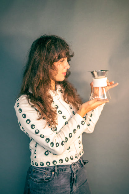 Woman holding a clear glass server with white silicone collar and stainless steel mesh filter against a grey background.
